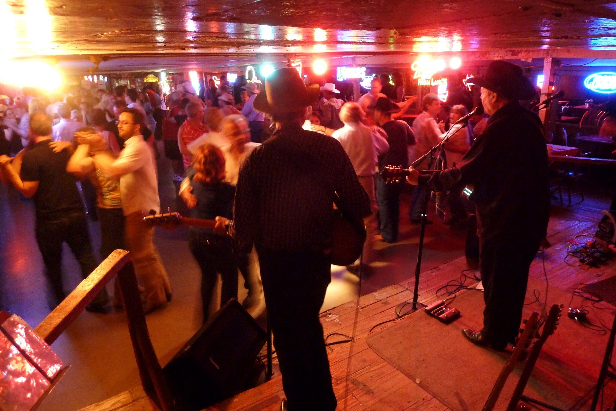 From stage, two musicians wearing black clothing and cowboy hats face a crowd dancing under glowing lights.