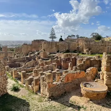 Excavated stone foundations and rows of low square pillars across a hilltop, a round millstone in front and a hazy plain beyond.