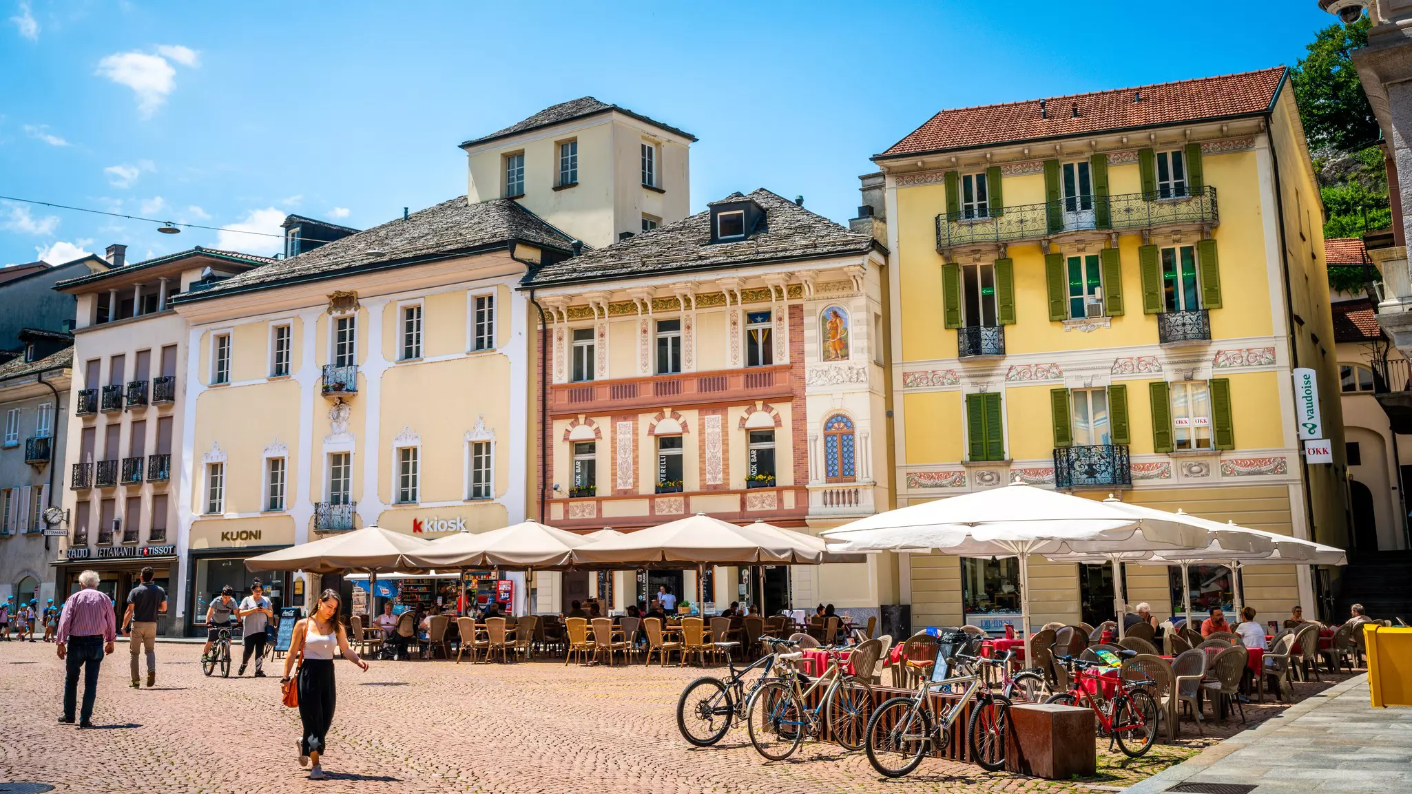People sit at outdoor cafes under umbrellas and walk through a plaza in Switzerland on a sunny day.