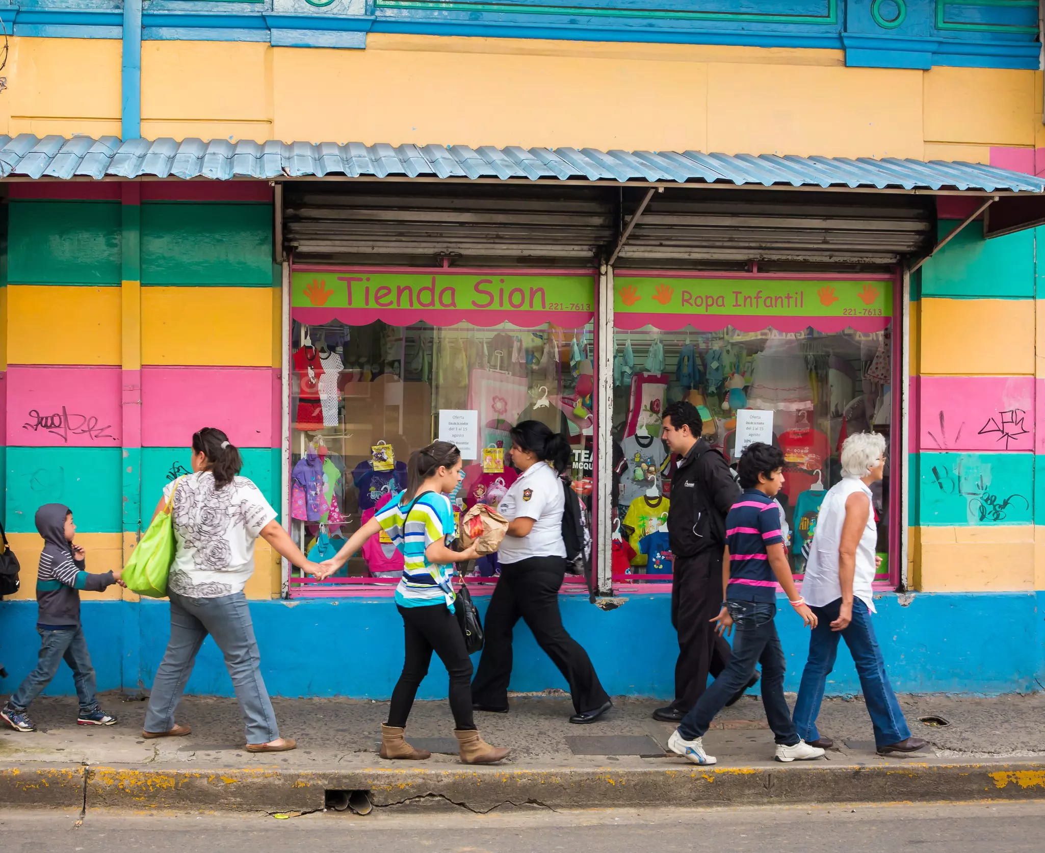 People walk in front of a shop in a city; the shop occupies a building with a facade painted in color-blocked stripes.