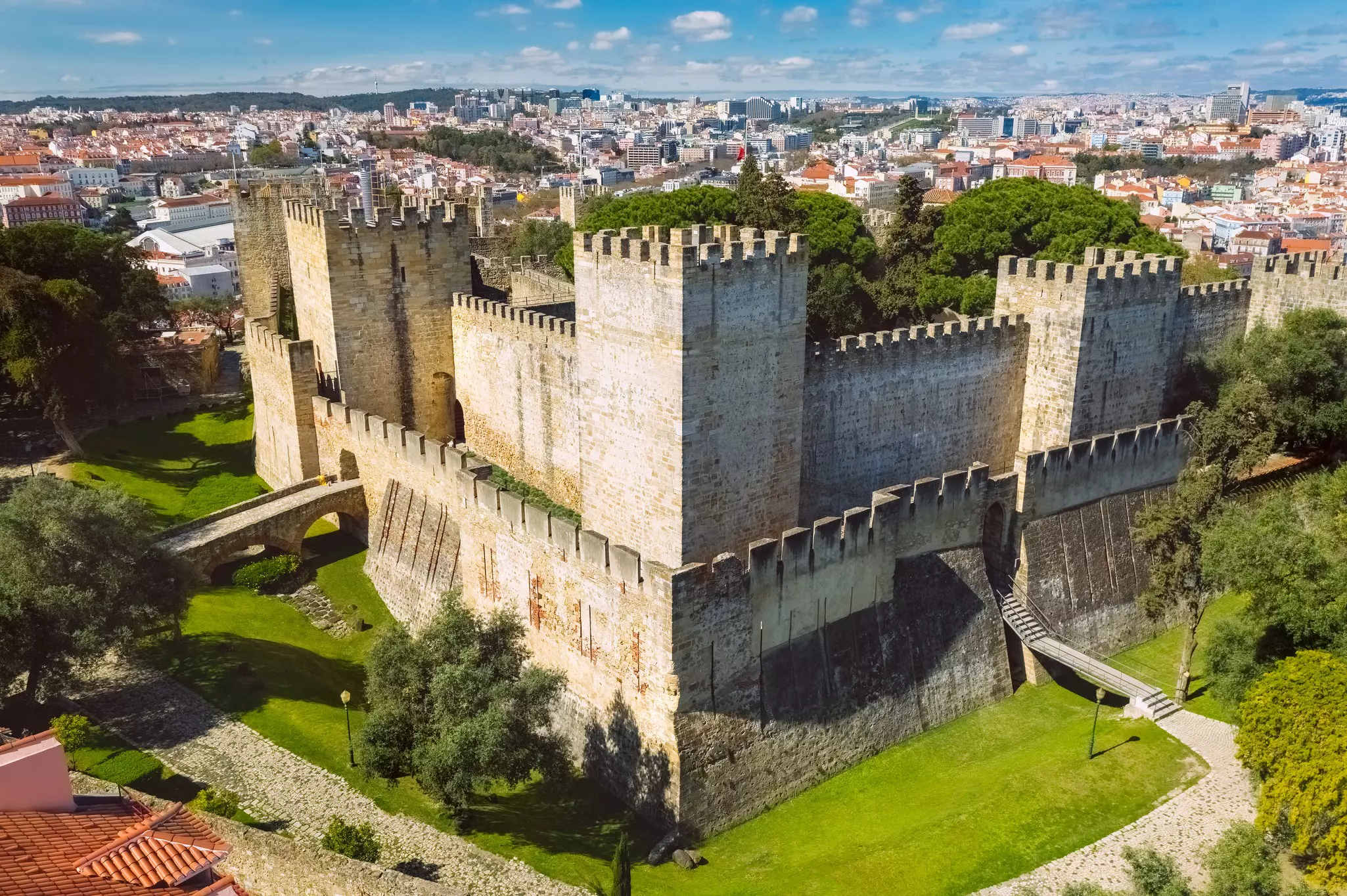 A wide view of a huge, restored hilltop castle with crenellated walls and towers. A large city is seen sprawling in the distance.