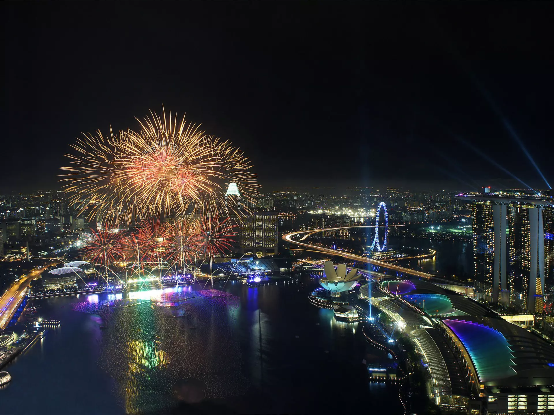 An aerial view of the Singapore NYE firework display. With a series of bright fireworks lighting up the sky above the city's skyscrapers.