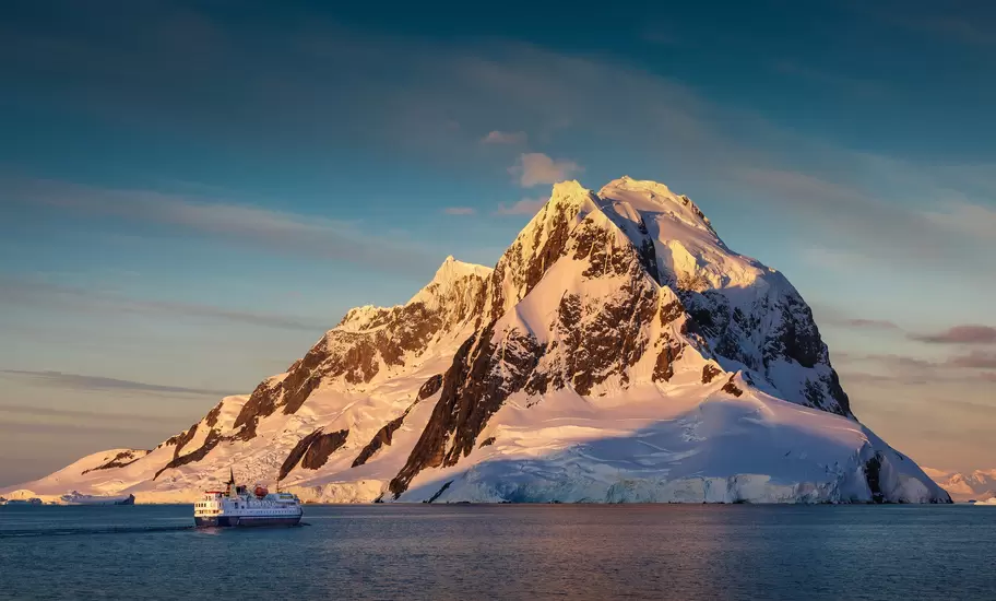 Antarctica: small expedition ship cruising beneath a massive snow-and-rock peak lit gold by the low sun