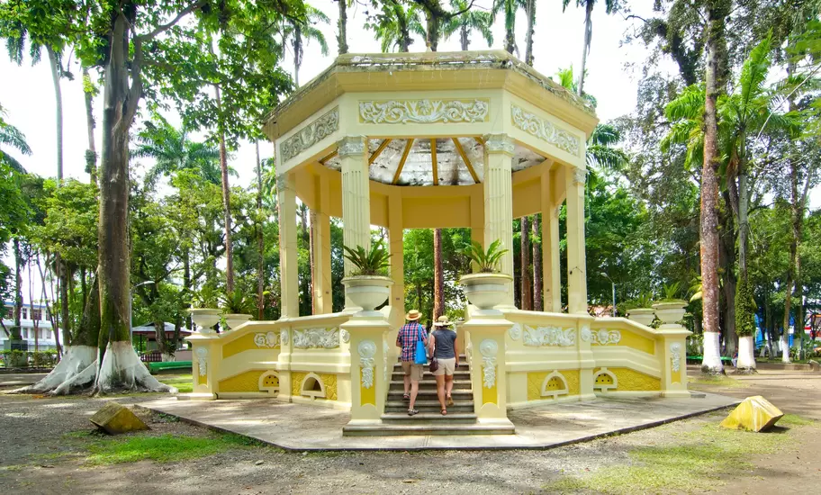 Two visitors climb the steps of an ornate yellow octagonal bandstand among tall palms and trees in a park