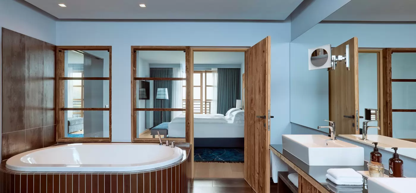 A luxurious hotel bathroom with a huge tub and a guestroom through the glass doors behind: COMO Hotels & Resorts, the Dolomites, Italy