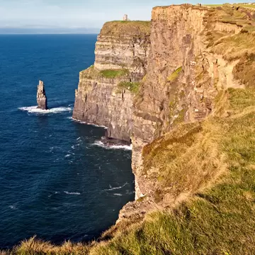 Sheer layered sea cliffs topped with grass and a small stone tower, a rock stack in the deep blue Atlantic below, Clare.