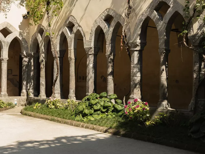 A stone courtyard with trees and plants surrounded by a historic stone building with arched designs along a rectangular path on a sunny day.