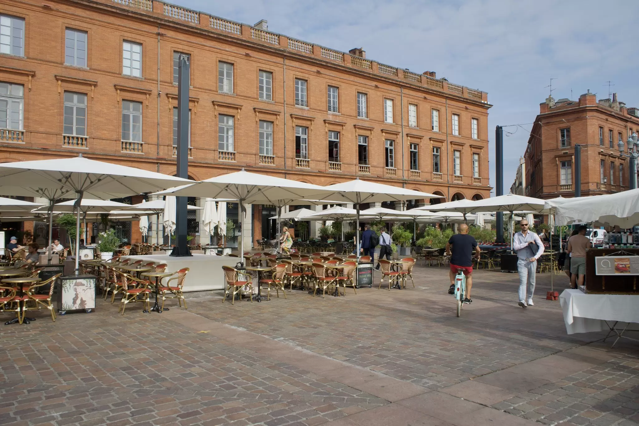 A series of restaurant tables with large white umbrellas line a square backed by a large red-brick building