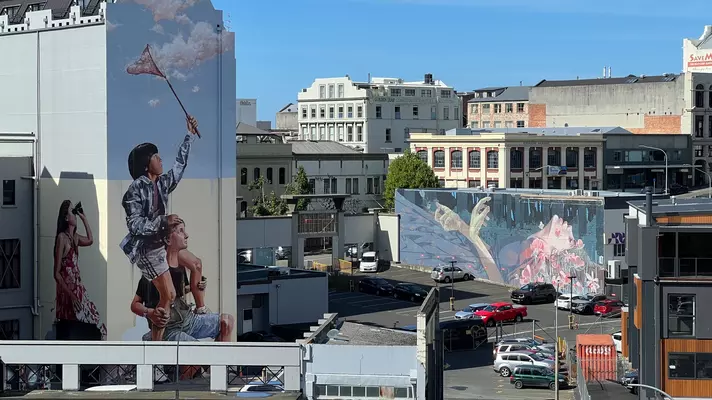 A cityscape with several large murals painted onto exterior walls of buildings, including a child on a man's shoulders waving a flag, and some hands in the air.