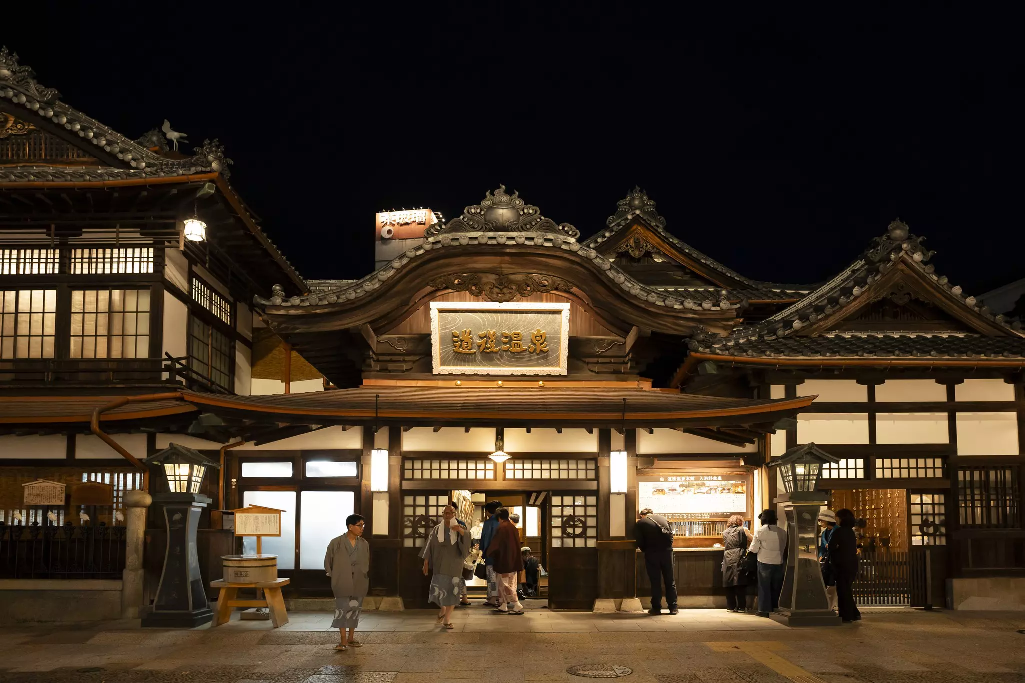 The exterior of a historic bath house in Japan at night. Patrons wander around wearing gray robes.