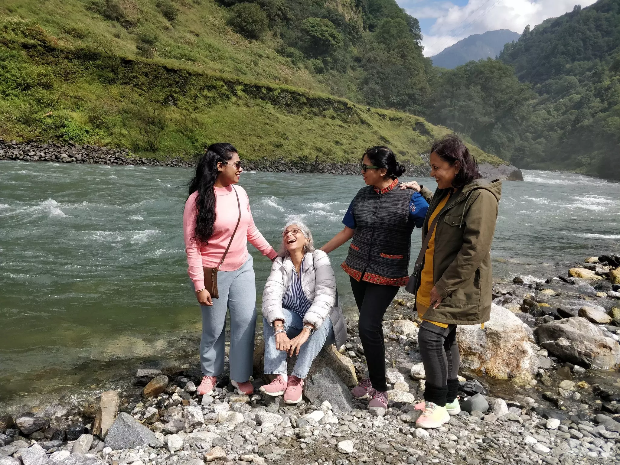A multi-generational group of Indian women pose for a photo on the rocks in front of a river with hills in the background.