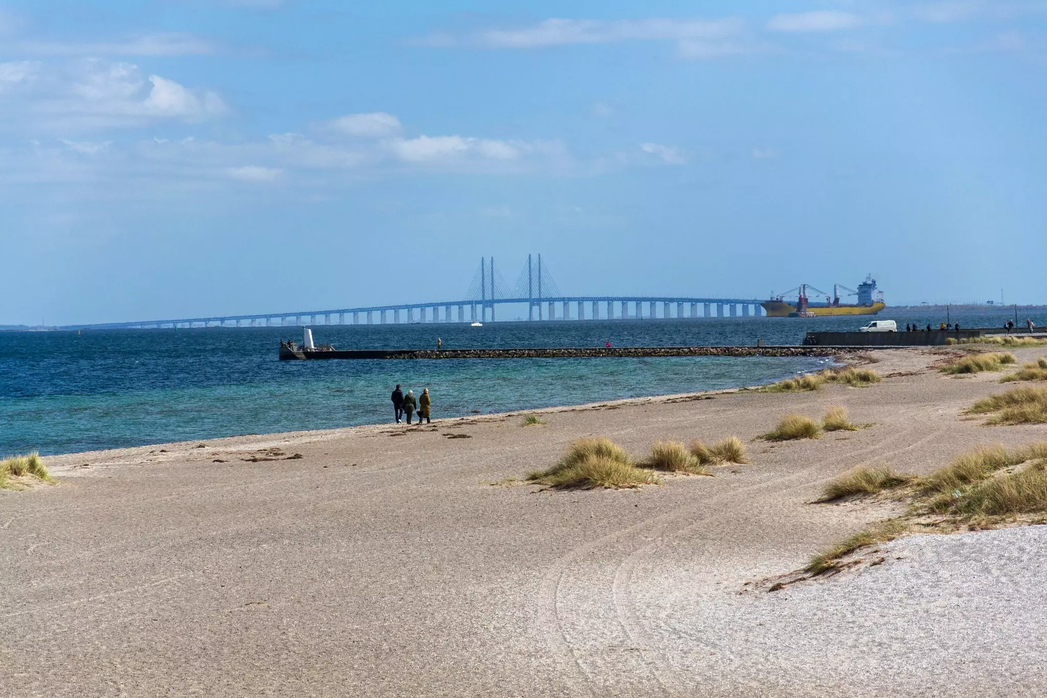 Three people walk on a sandy beach with tufts of grass; a bridge is on the horizon.