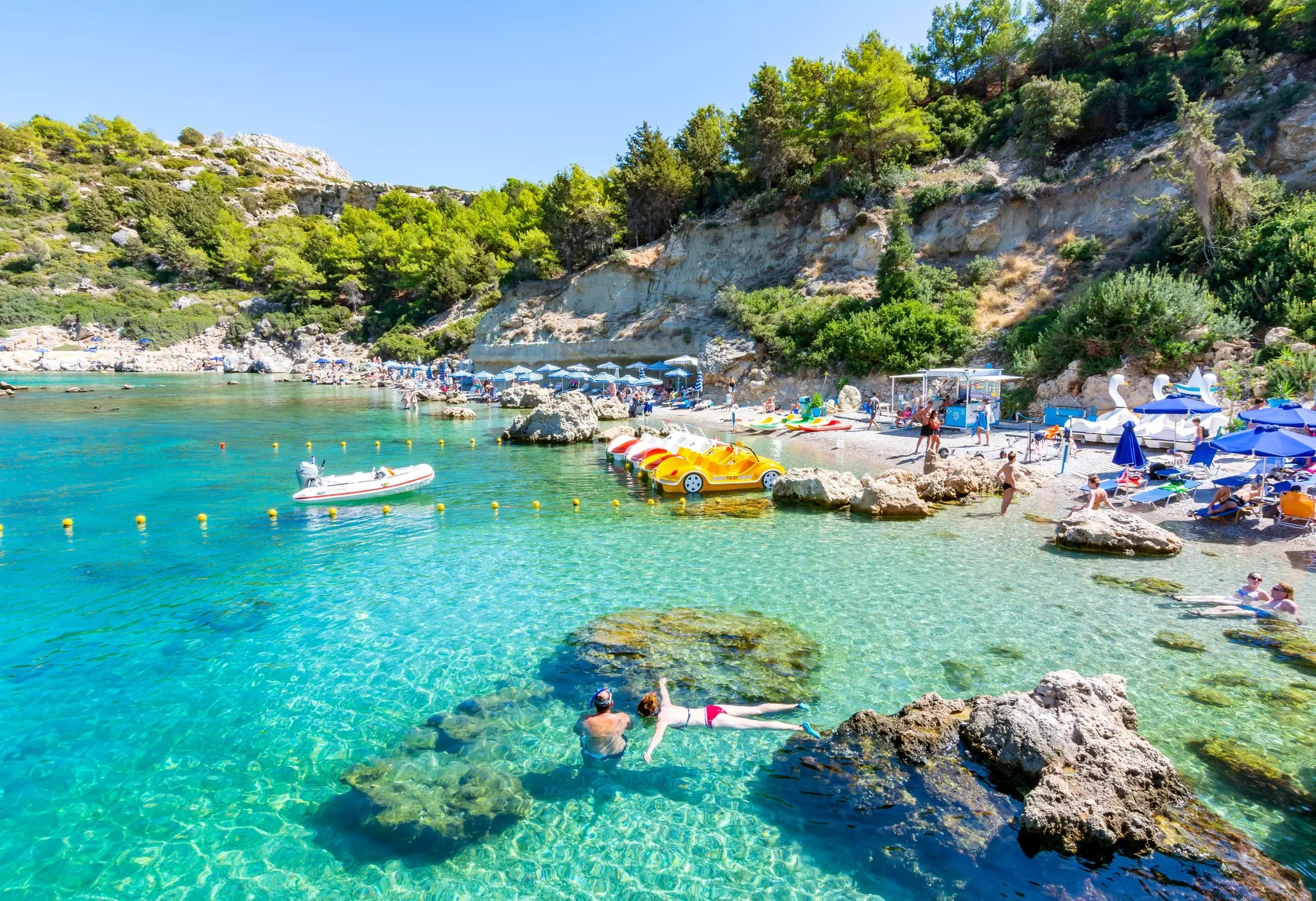People sunbathe under blue umbrellas on a beach with clear water