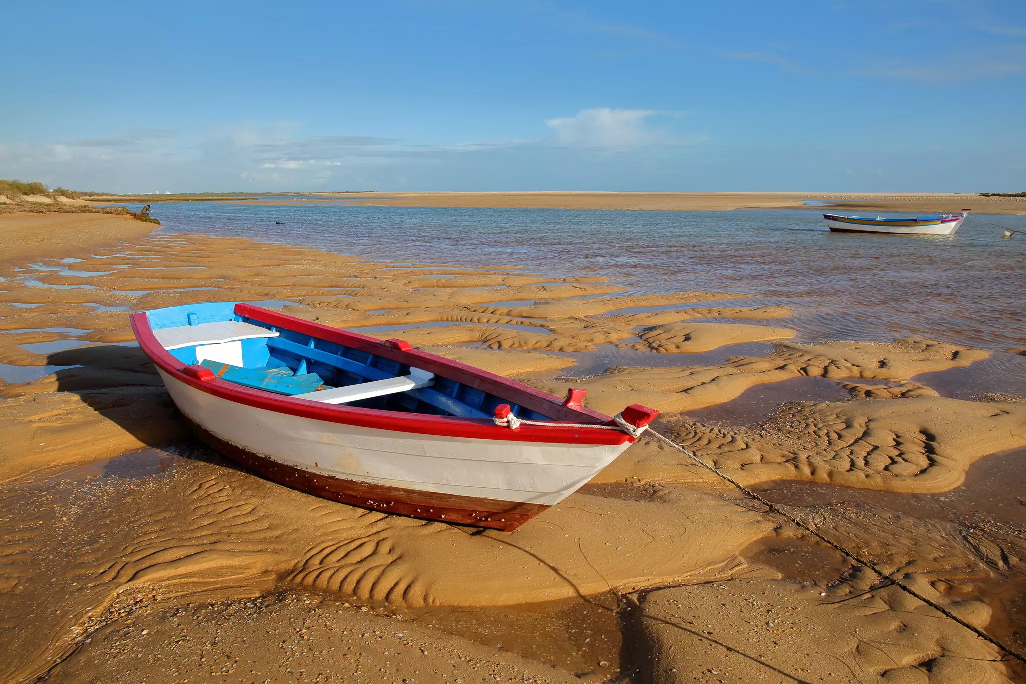Two boats, one in foreground and one in background, moored on a sandy spit of land near a low-tide ocean.