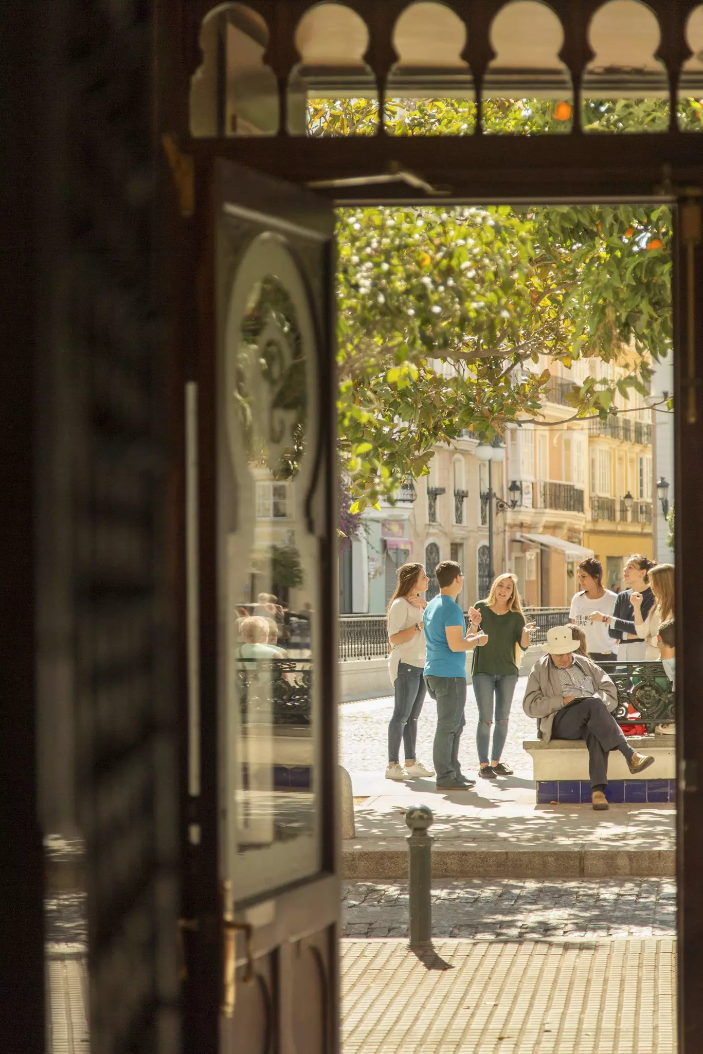 People sitting in a plaza overlooked by orange trees are viewed through an open doorway.