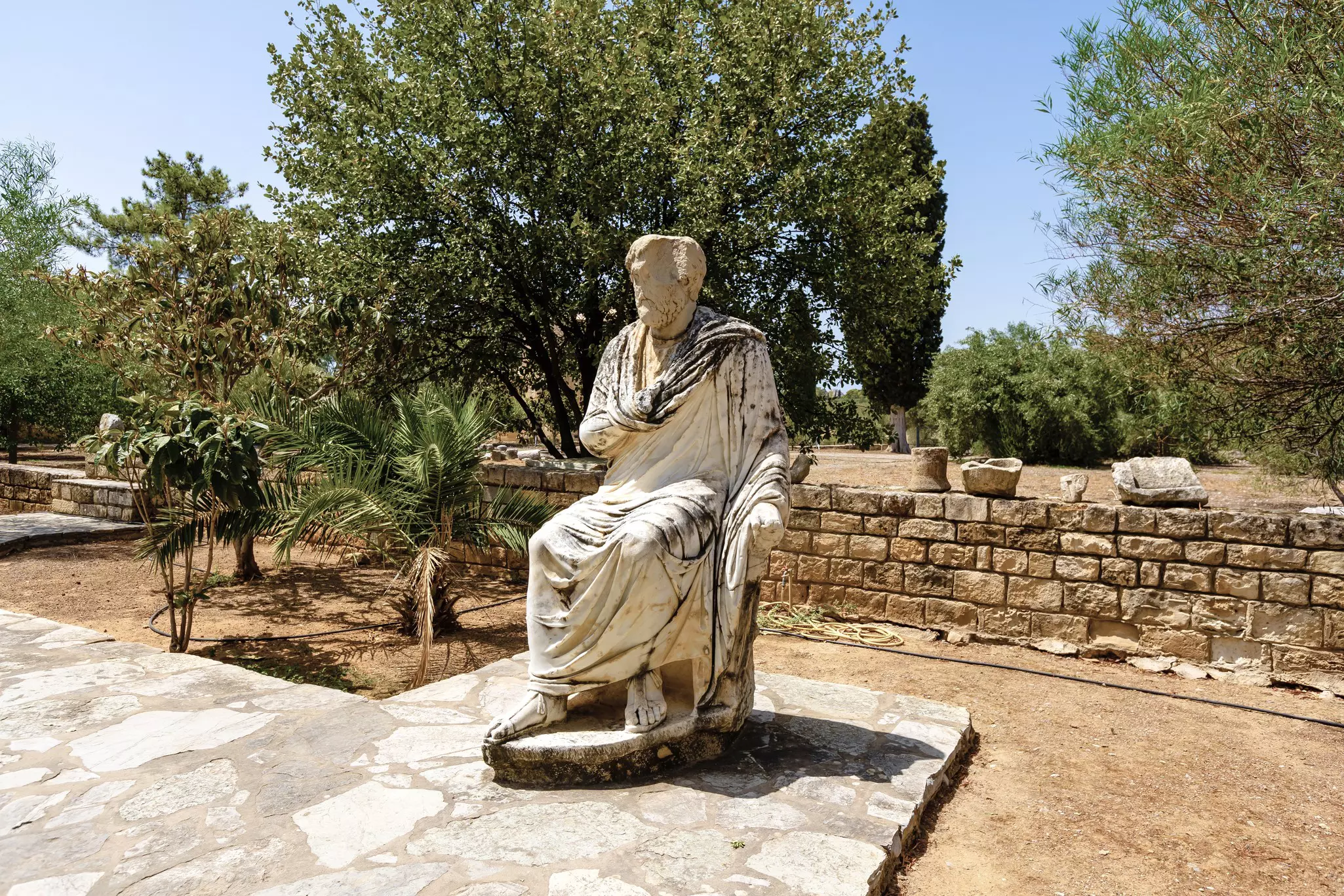 A full-scale stone sculpture depicts a seated figure in a toga at the outdoor archaeological site of Gortyna, Crete
