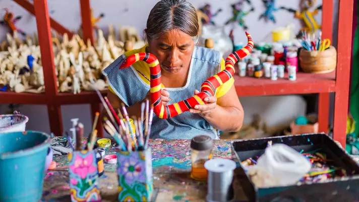 A woman painting a wooden snake red with yellow and black stripes.