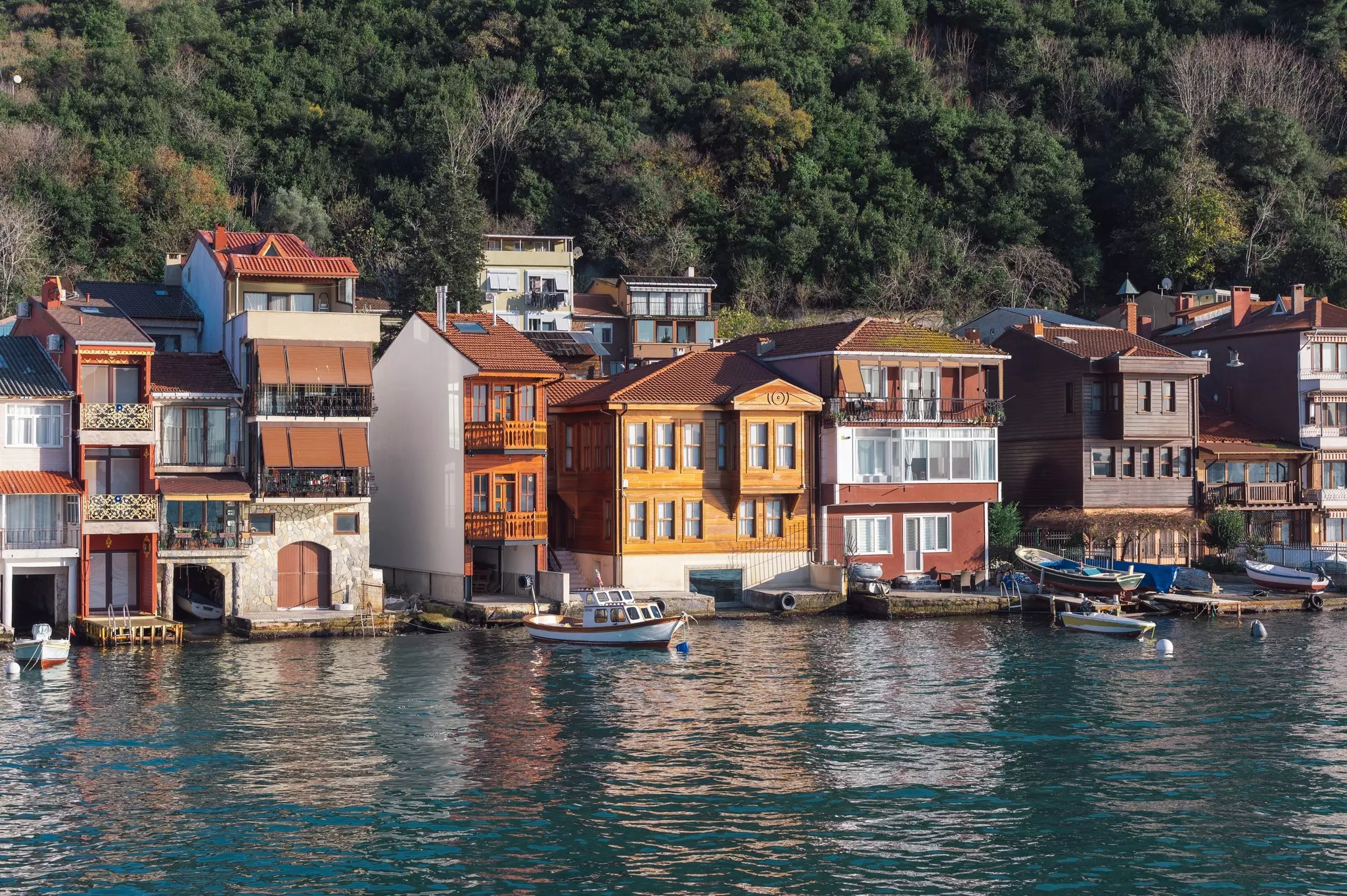 Water laps a row of four-story houses, with boots moored in front and a hilly forest behind.
