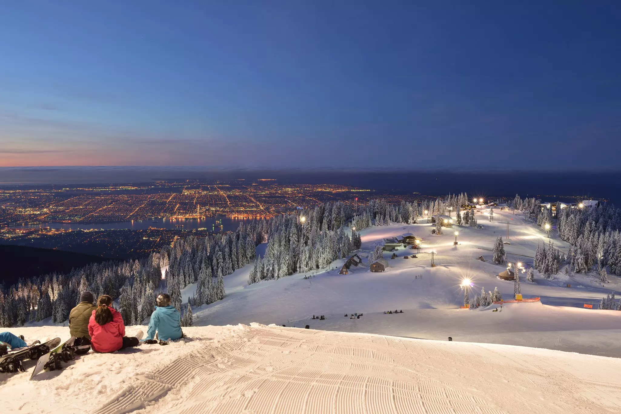 Three snowboarders sit at the top of a ski slope in the early morning just as dawn begins to break in the distance.
