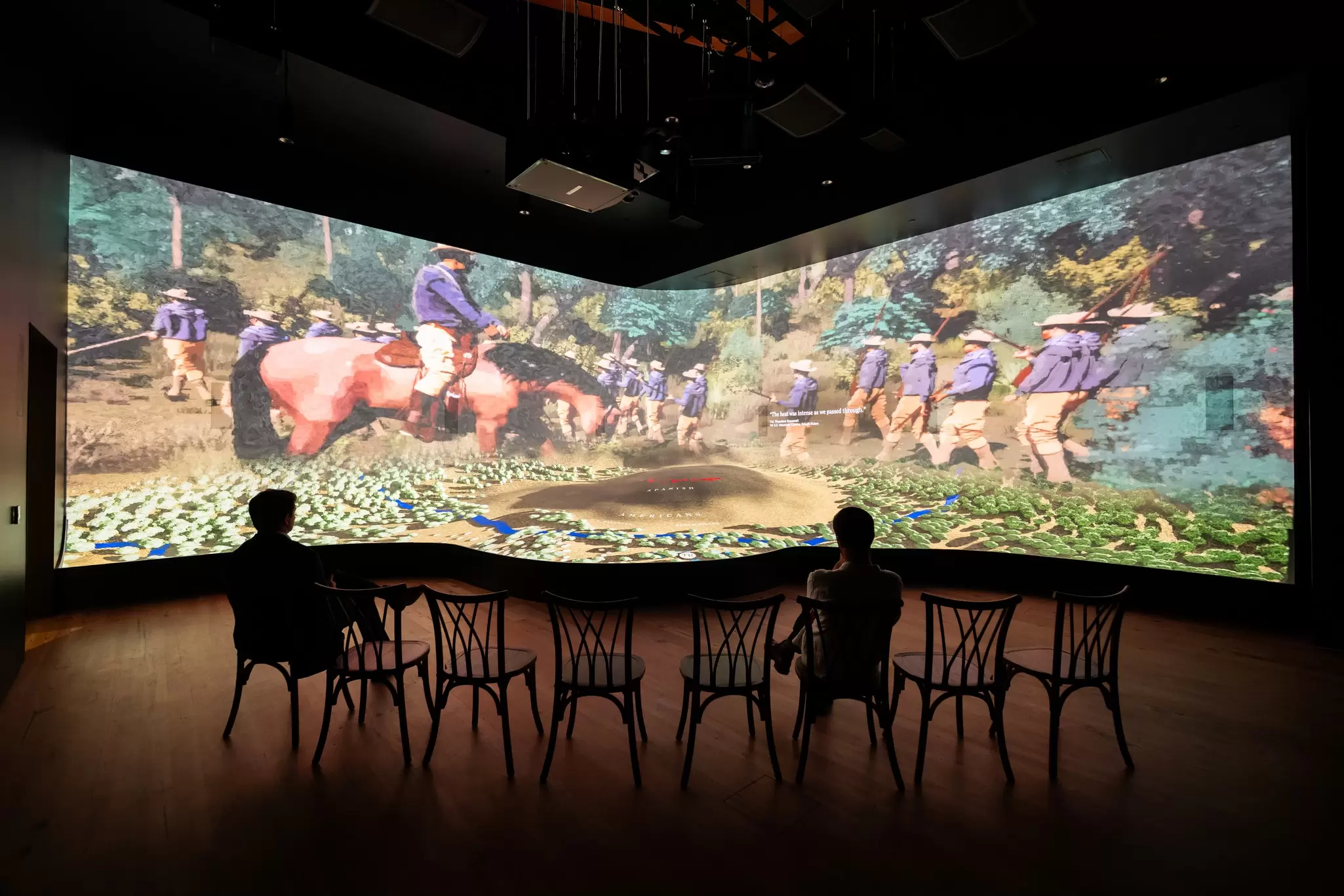 People sit in chairs while watching a video exhibit at the library.