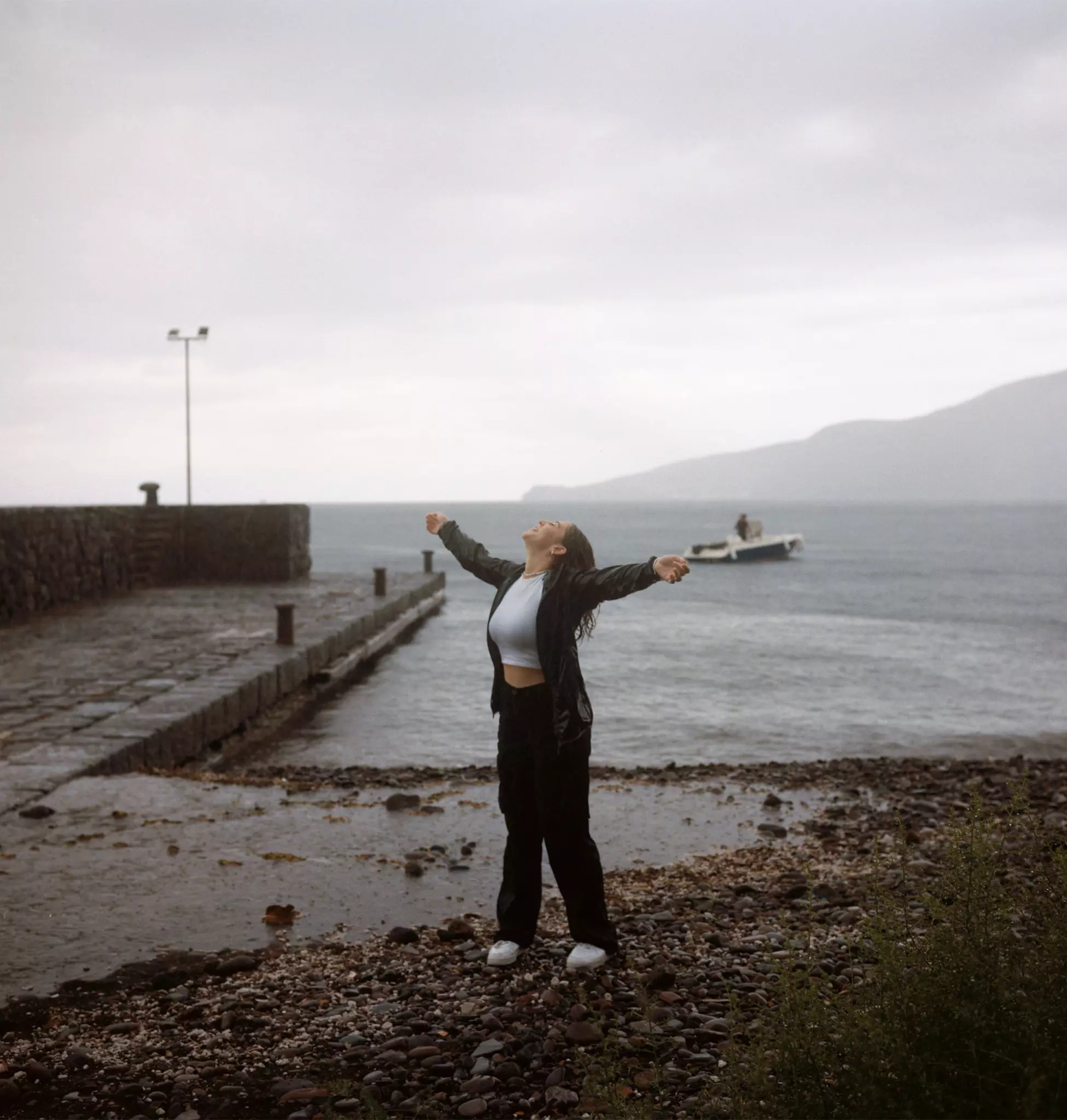 A young woman stands by a pier in a town on a rainy day and throws her arms back.