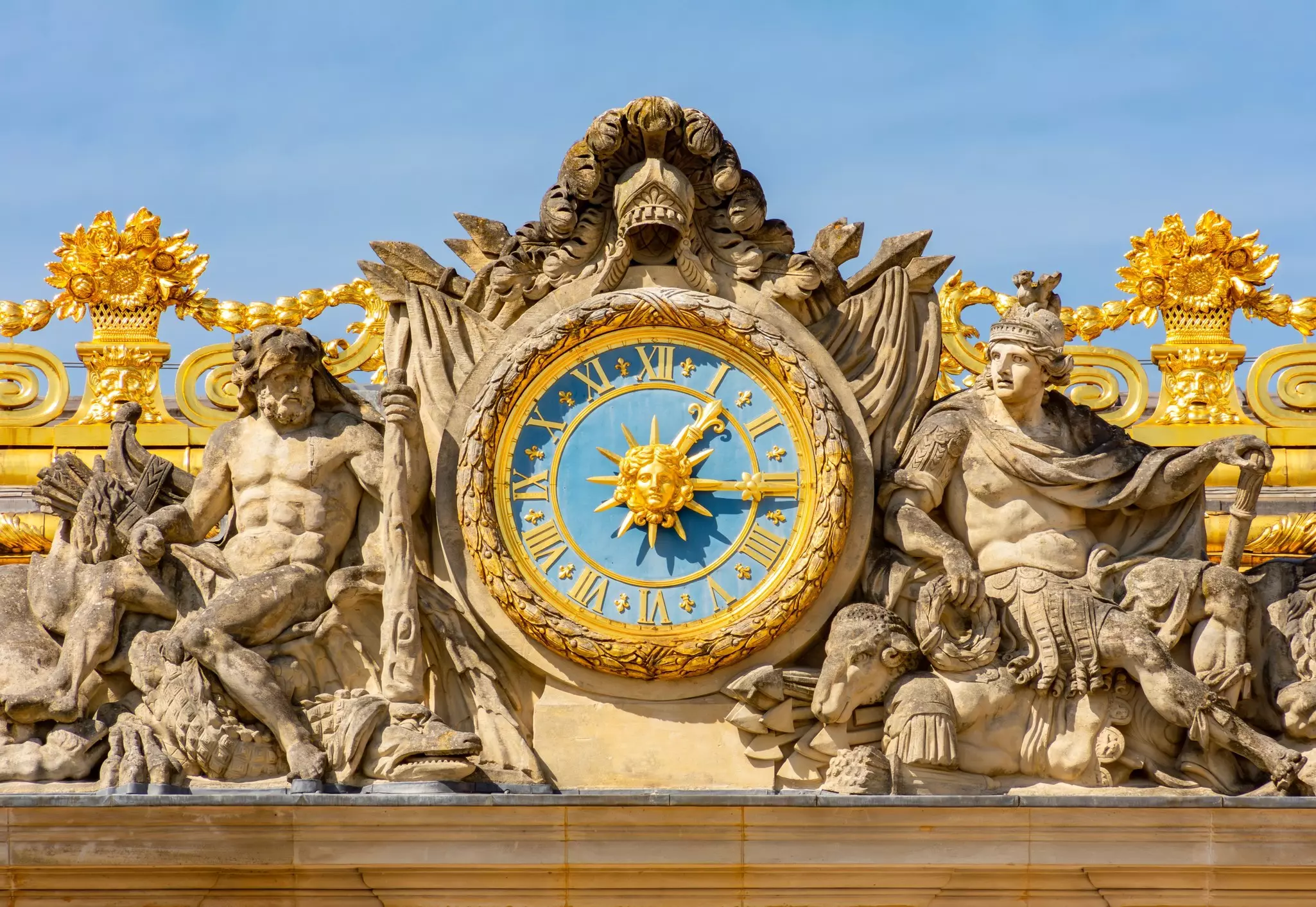 Figurative carvings on either side of a gold clock on a building facade.