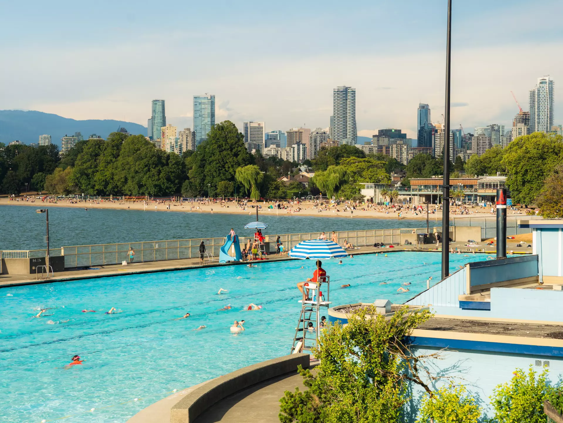 Swimmers at Kitsilano Pool in Vancouver watched over by a lifeguard. A distant beach is backed by high-rise buildings