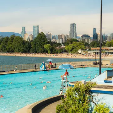 Swimmers at Kitsilano Pool in Vancouver watched over by a lifeguard. A distant beach is backed by high-rise buildings