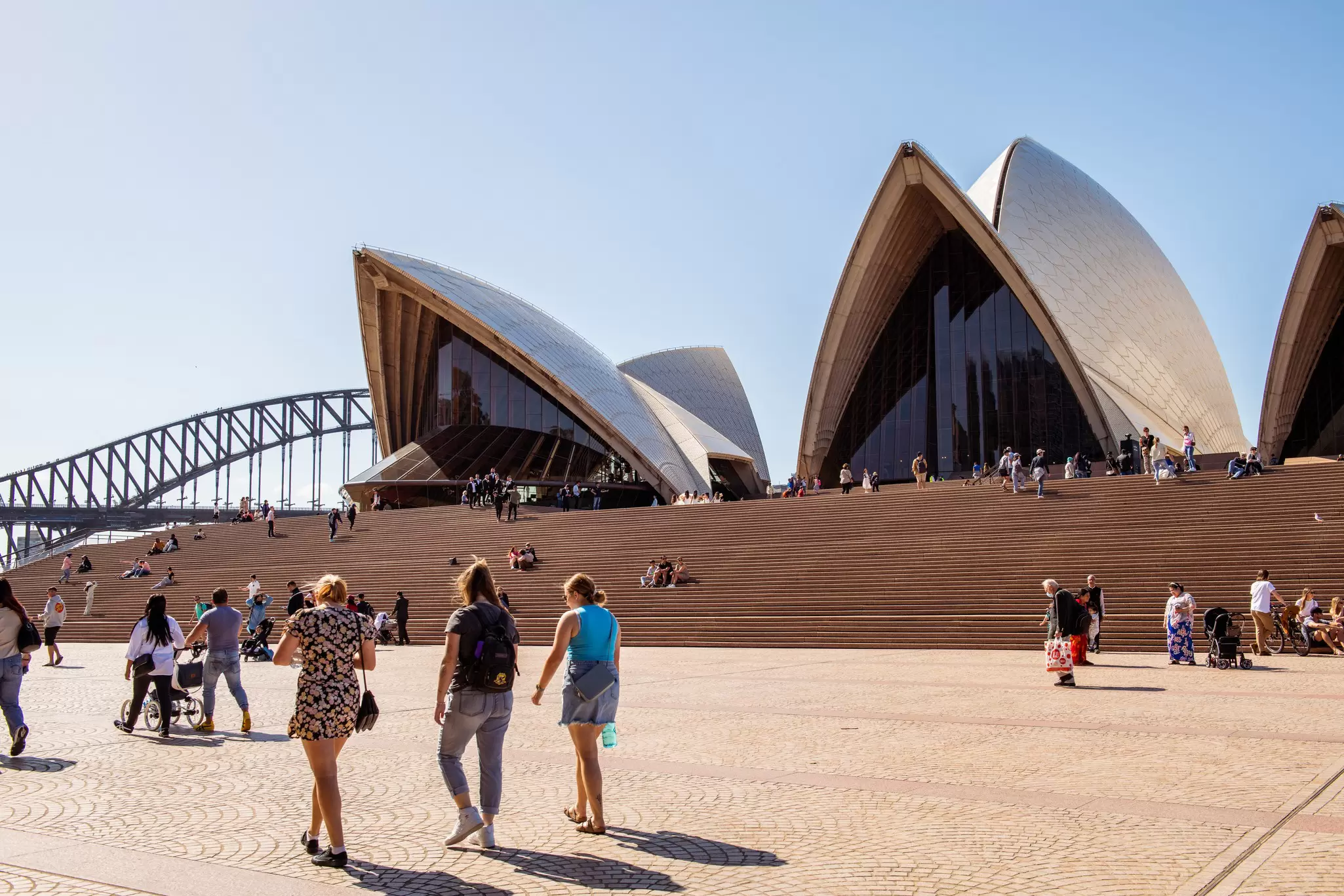 People walk in front of the Sydney Opera House on a sunny day in Sydney, Australia.