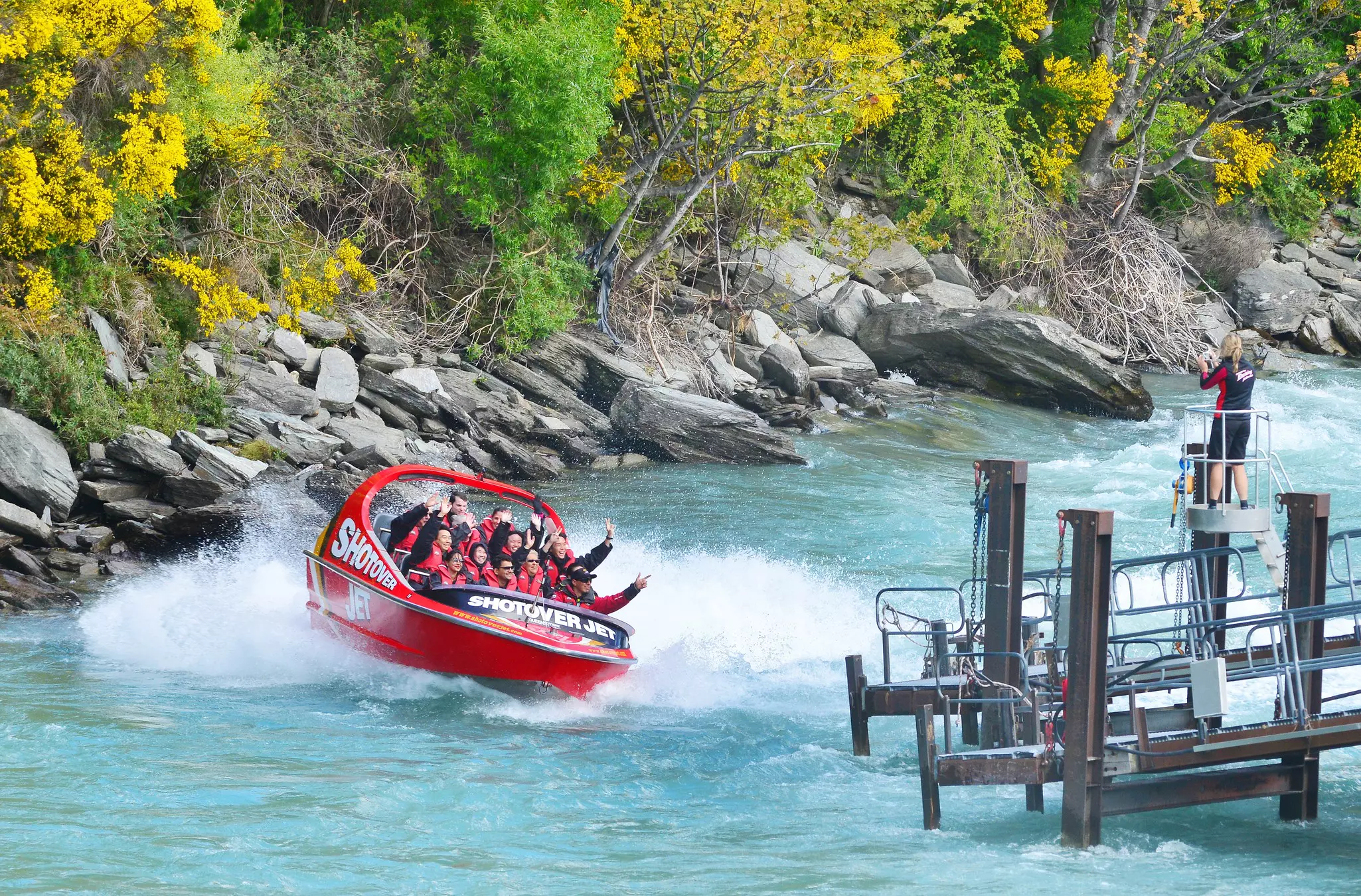 Passengers on a red speed boat out on a river all smile at a photographer standing on a dock.