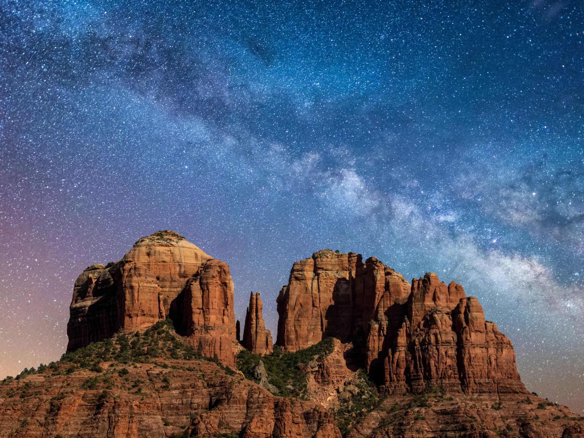 A towering reddish-brown rock formation stands against the night sky, illuminated by the stars of the Milky Way
