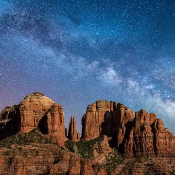 A towering reddish-brown rock formation stands against the night sky, illuminated by the stars of the Milky Way