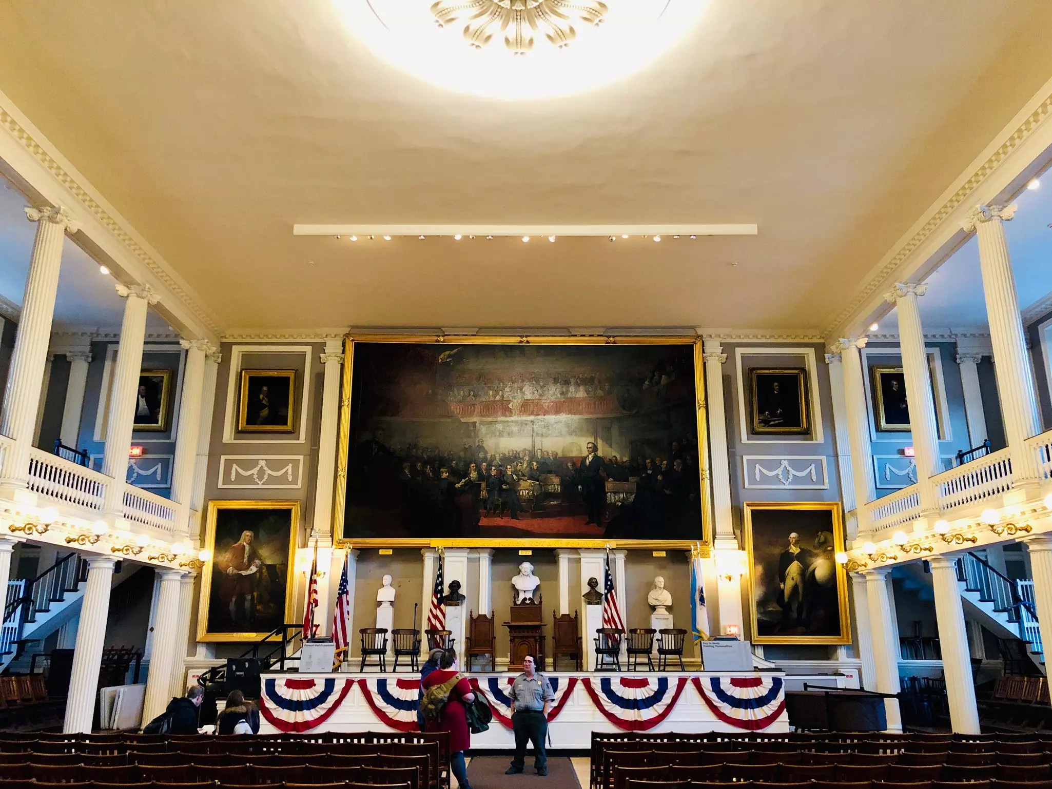 A person gives a tour of a historic meeting hall, lined with columns, with large paintings hanging on the wall at the front of the hall.
