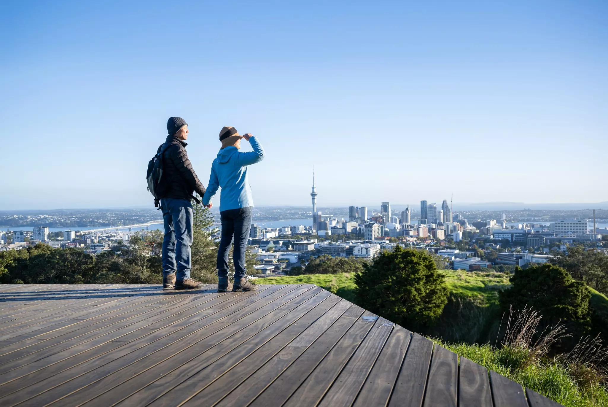 Couple standing on boardwalk at Mt Eden summit. Sky Tower and Auckland Harbour Bridge in the distance. Auckland., License Type: media, Download Time: 2025-03-20T20:27:15.000Z, User: mvm_lonelyplanet, Editorial: false, purchase_order: 56530 - Guidebooks, job: New Zealand 22, client: Global Publishing-WIP, other: Virginia Moreno