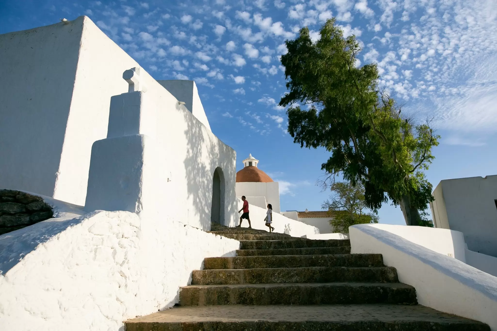 Two figures wander up a path towards a low-rise white-washed church in Santa Eulària des Riu