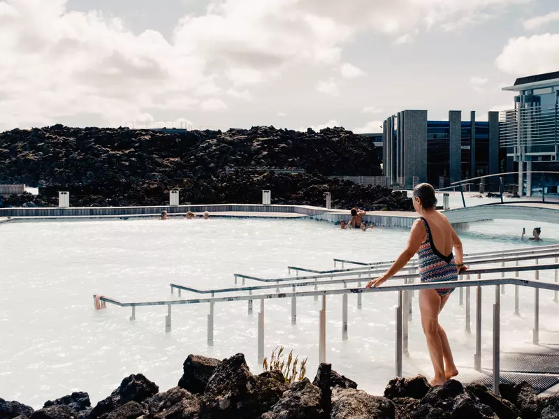 A woman in a bathsuit walks down a ramp toward a large soaking pool with a glass-sided building to the right and a rocky hillside in the distance on an overcast day.