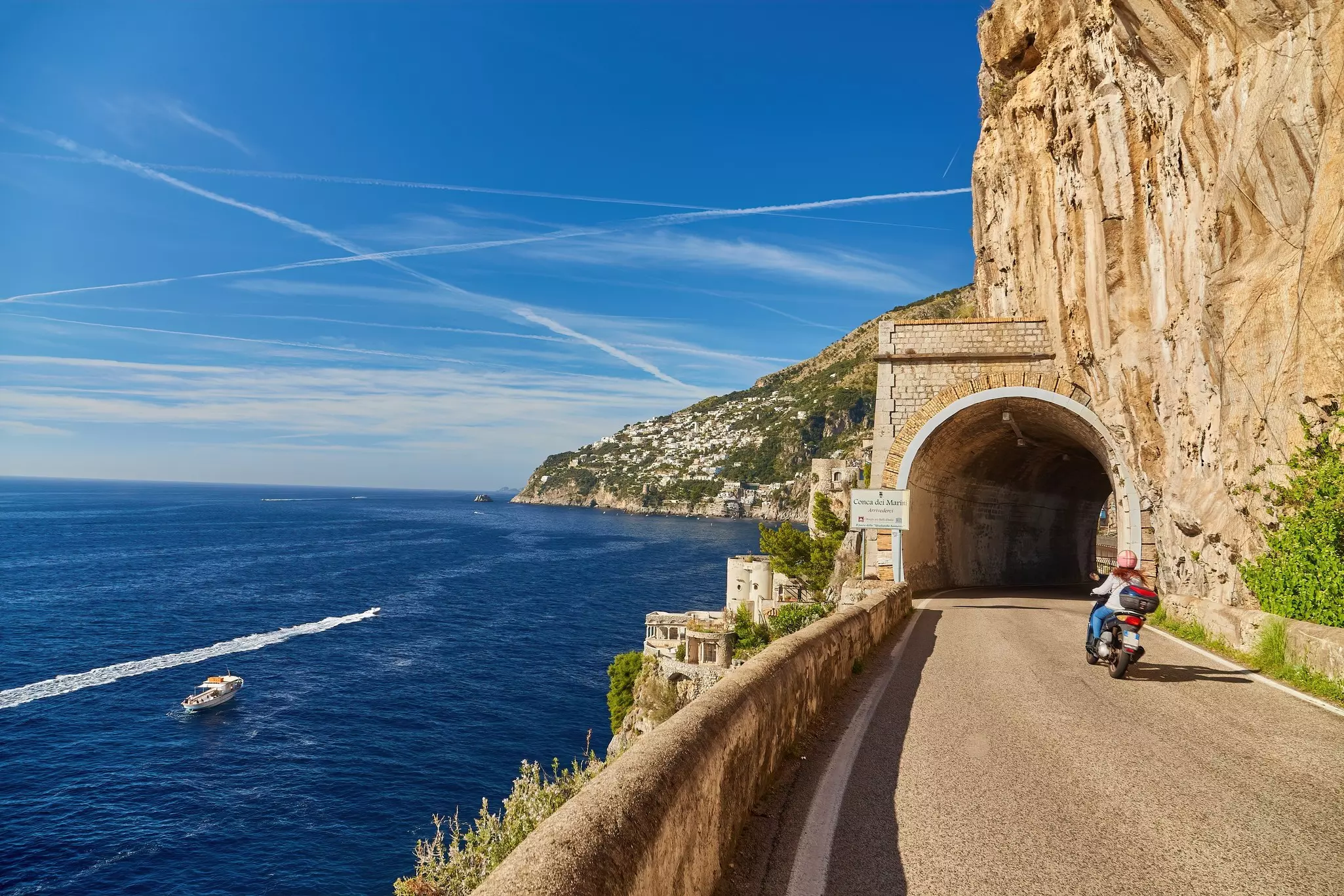 A scooter approaches a tunnel on a road hugging the cliffs of a coast. Boats are seen in the sea below the road.
