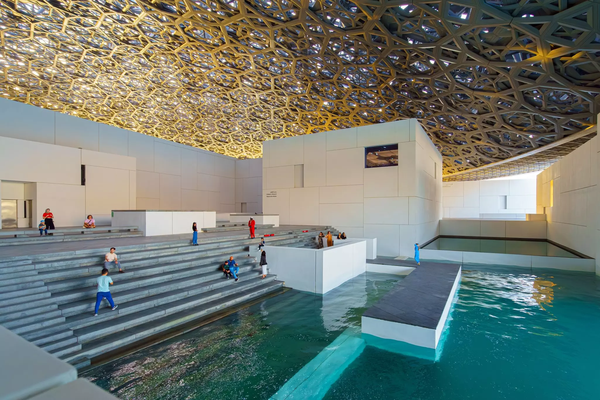 A museum with steps down to a vast indoor water feature and a detailed mesh ceiling.