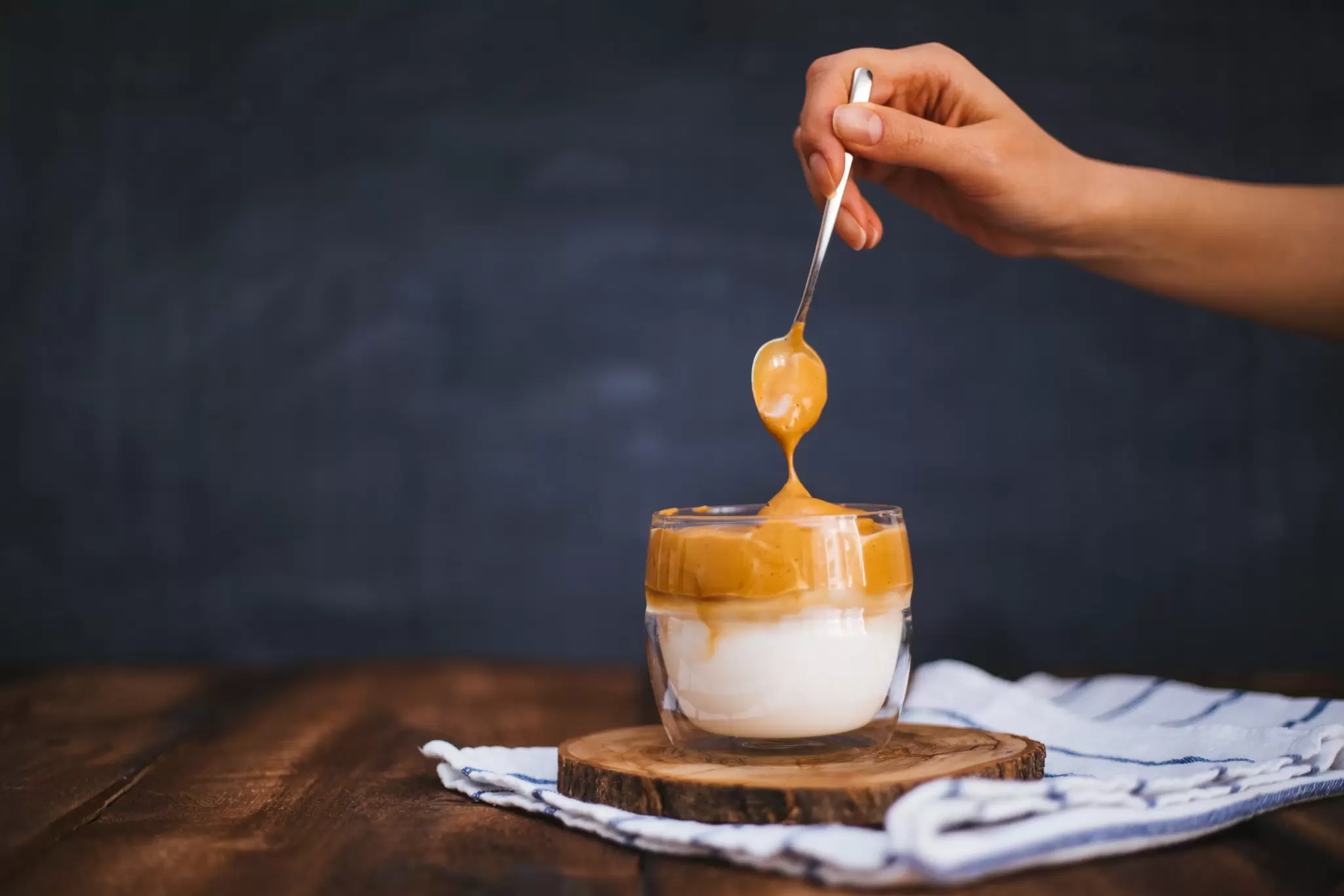 Hand spooning whipped caramel-colored dalgona coffee foam over milk in a small glass on a wooden board