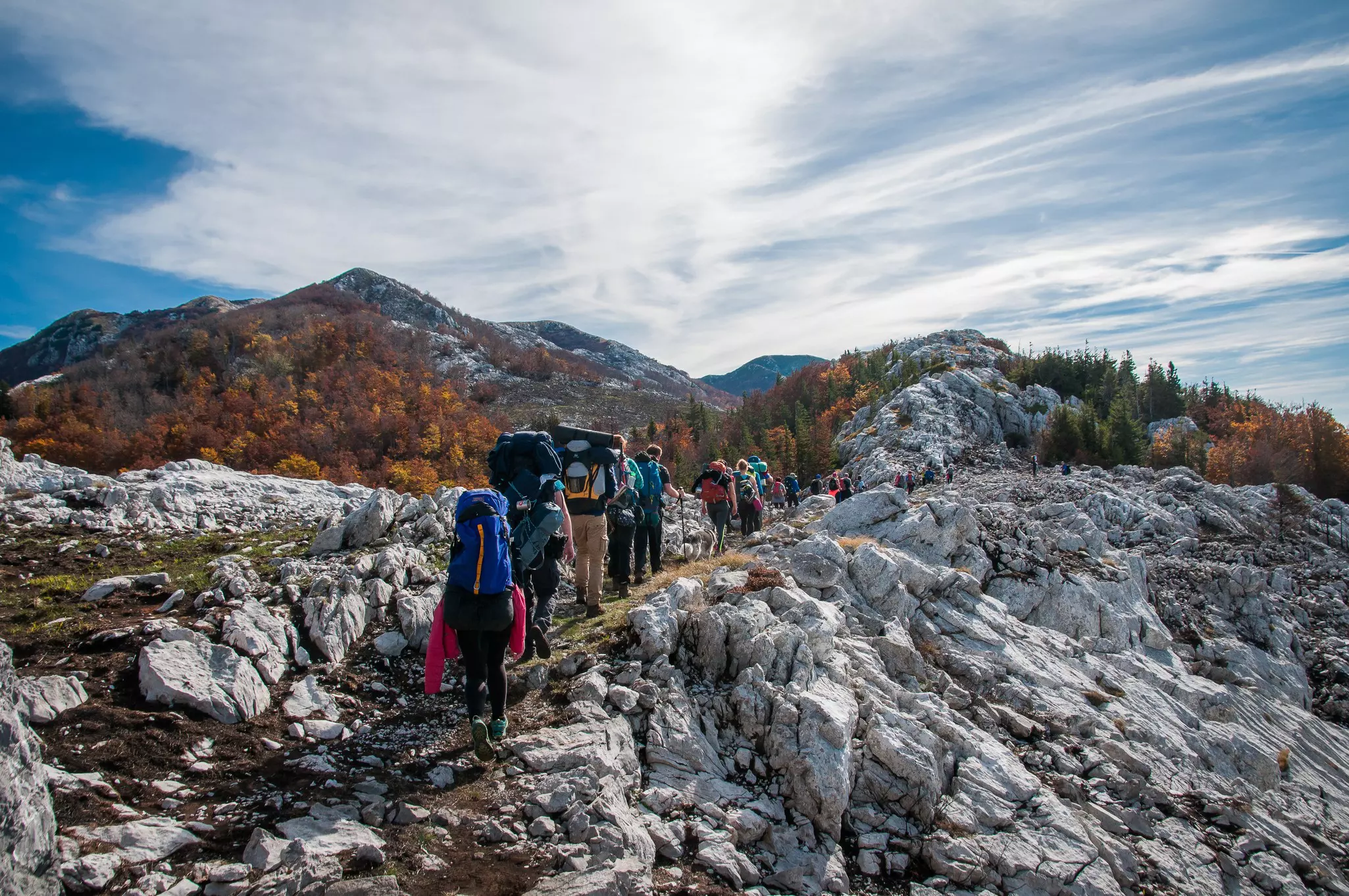 A row of hikers laden with heavy backpacks follow a rocky mountain trail