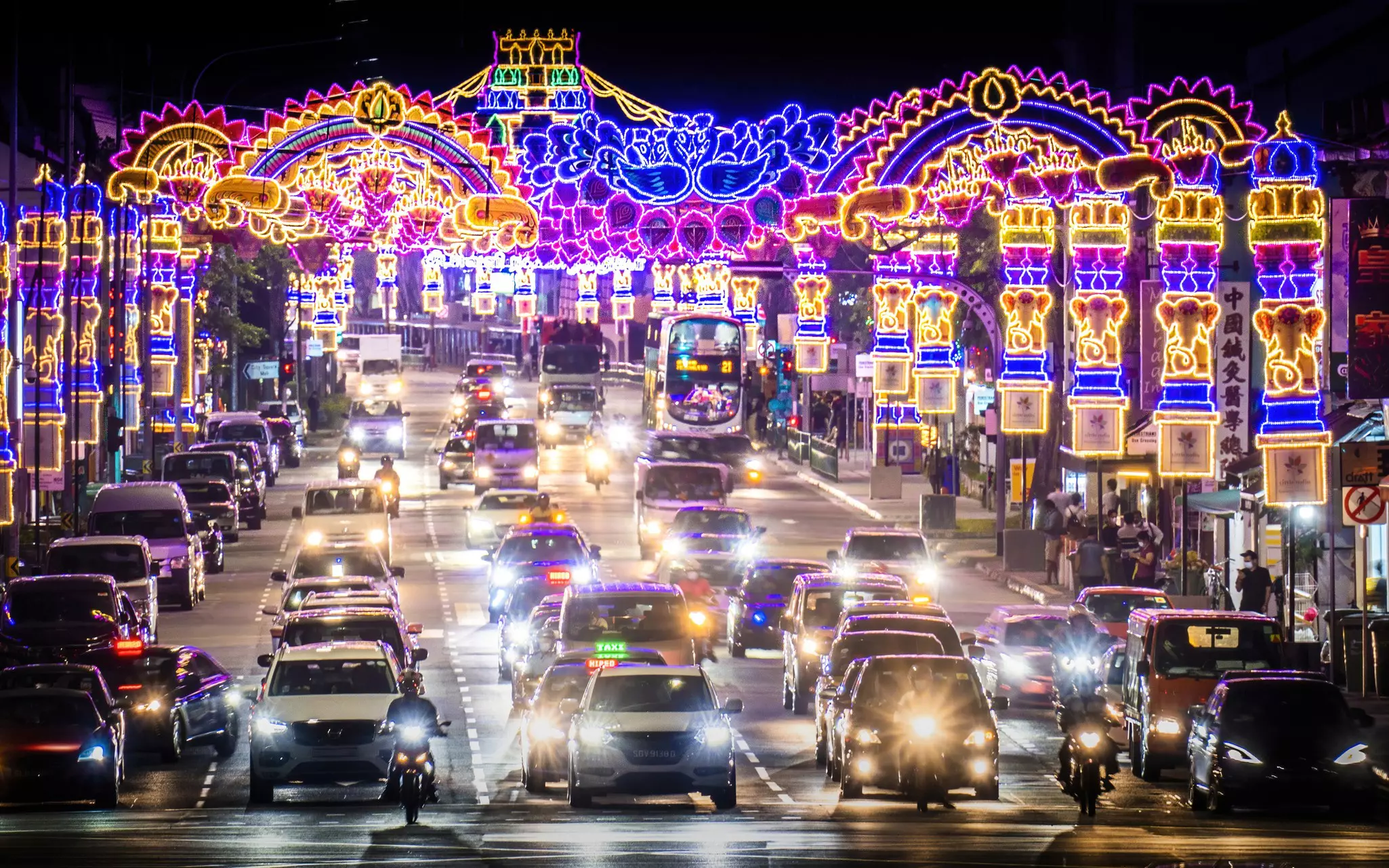 A row of cars in a street is seen at night, with brightly lit decorations festooning the street.