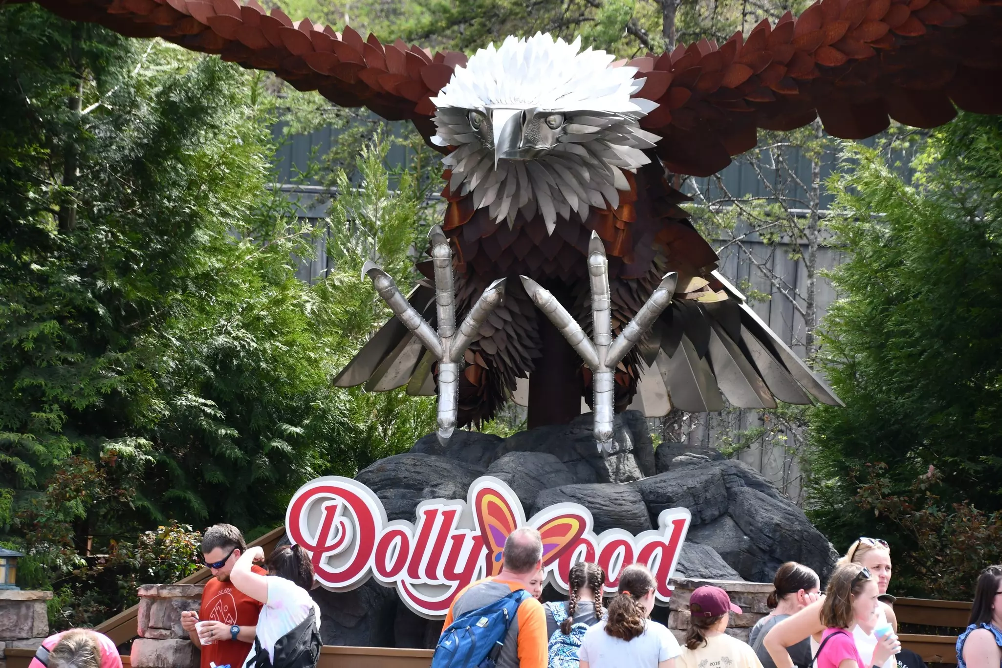 People at an amusement park stand in front of a giant wooden sculpture of a bald eagle.