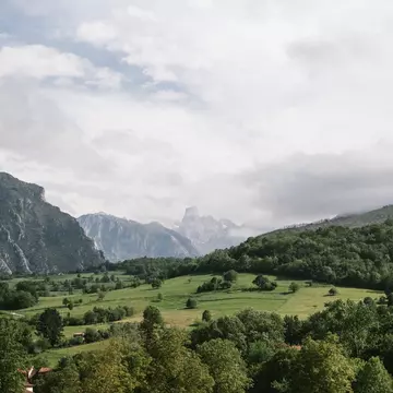 A green field surrounded by trees with rocky peaks in the background under cloudy skies.