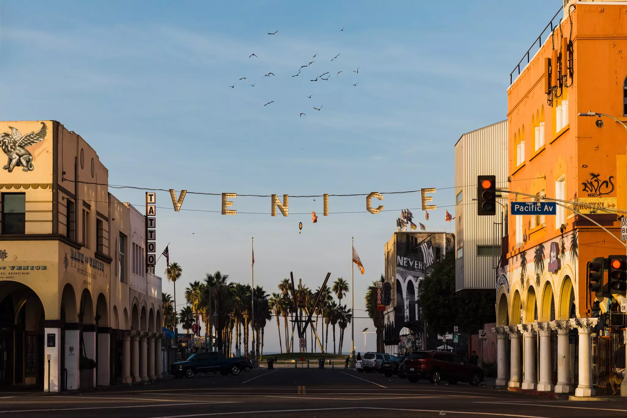 Early morning sun on a street lined with arched buildings, palm trees in the background and the word Venice hanging along a wire stretched across the street.