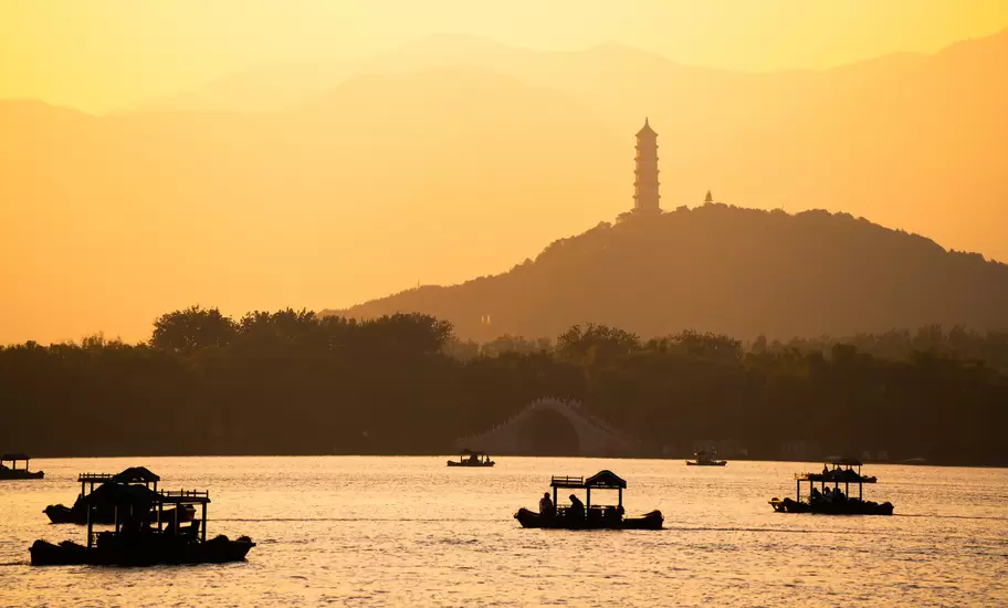 Silhouetted pleasure boats on a calm lake at sunset, with a hilltop pagoda and arched bridge in golden haze