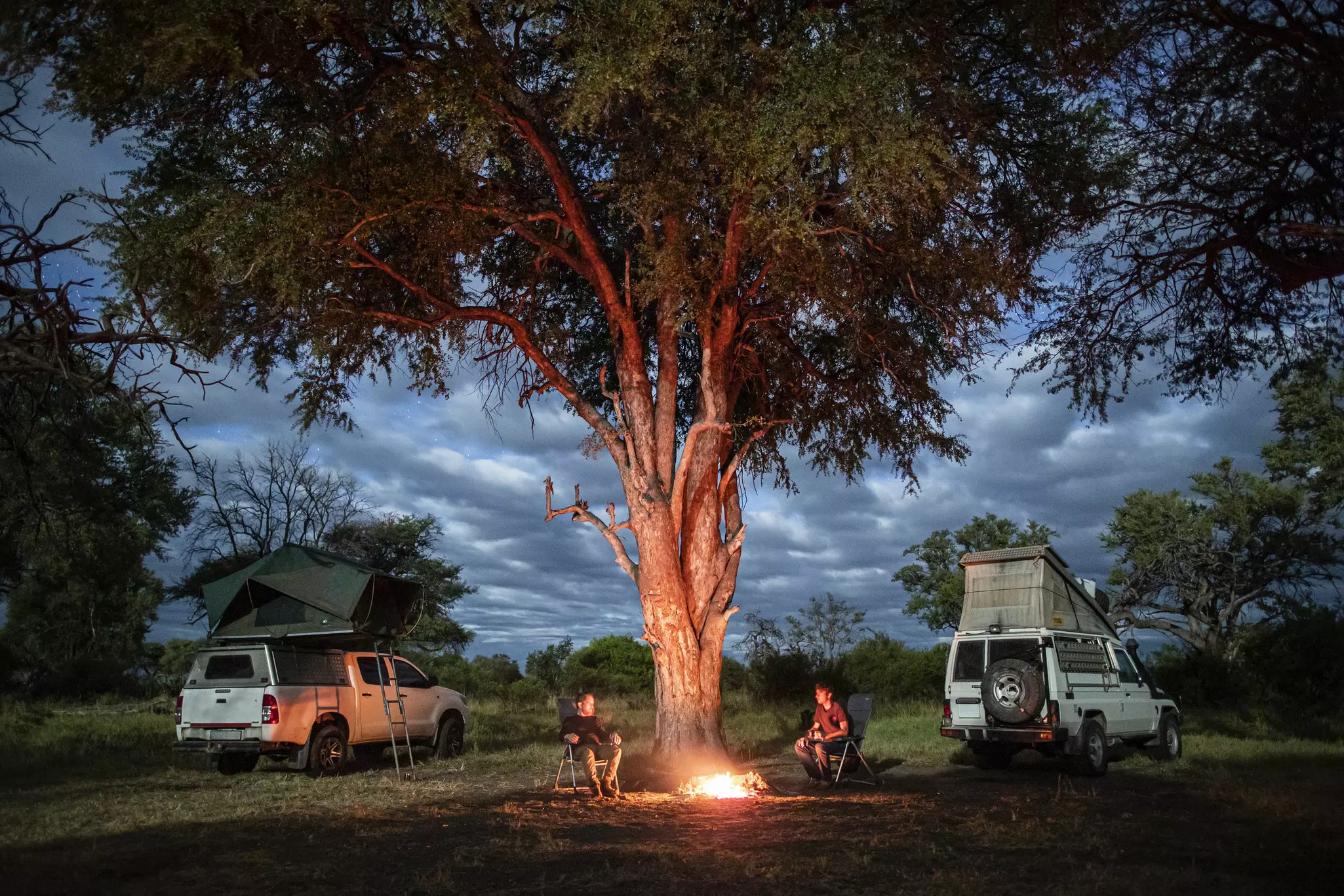 Two men sit near a fire beneath a tree in the African bush next to their camping 4WD cars, which have the roof tents popped up