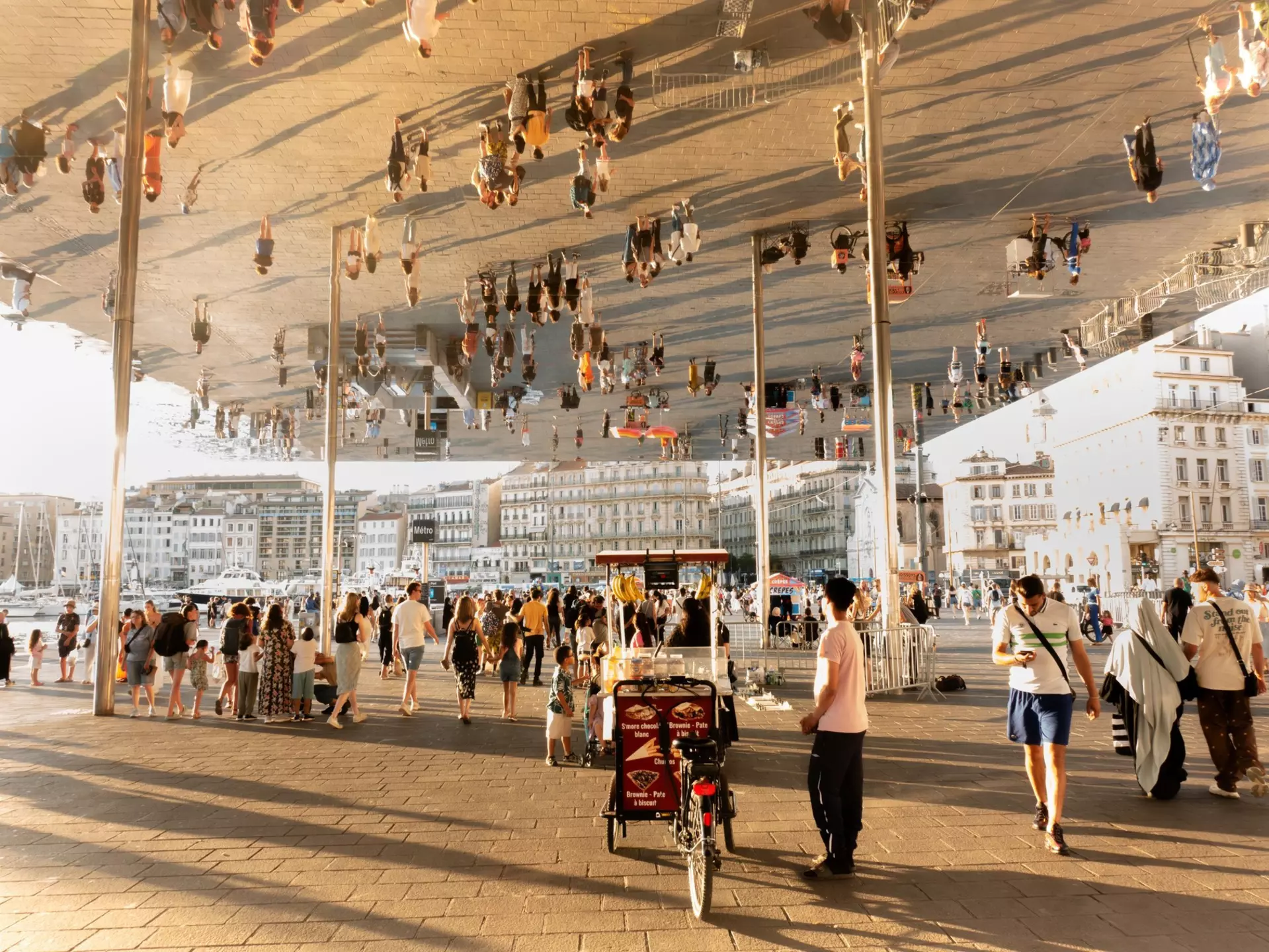 People crowd under a reflective pavilion in a city square by the port.