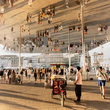People crowd under a reflective pavilion in a city square by the port.
