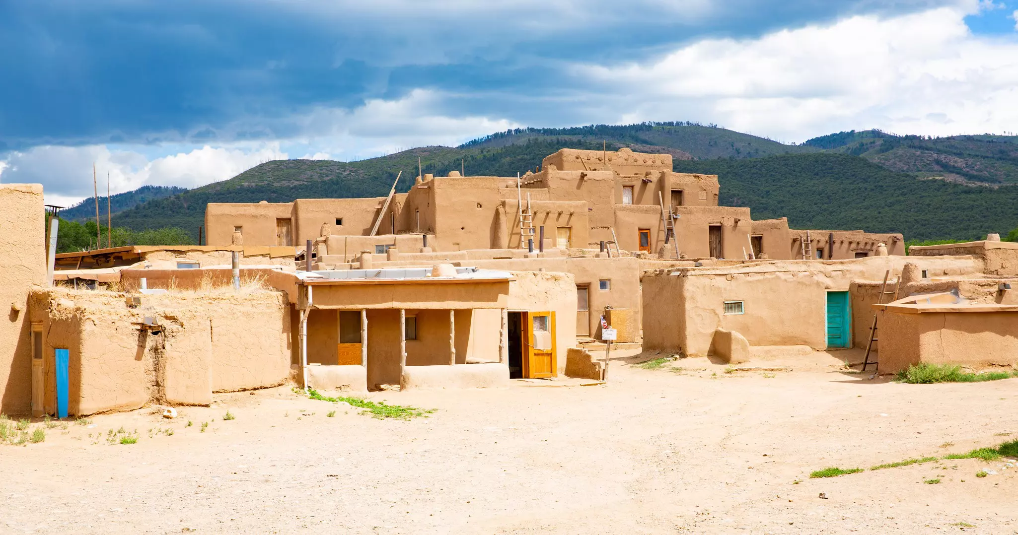 A series of pueblo dwellings atop dry earth with forested mountains in the distance on a partly sunny day.