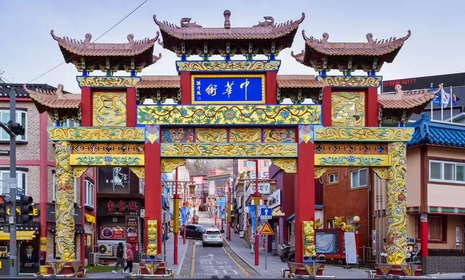 Red, gold and green Chinese-style gate with tiled roofs over a street lined with shops in Incheon Chinatown, South Korea.